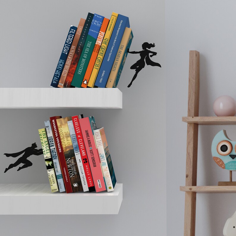 May include: Two white shelves with books on them. The top shelf has a superhero silhouette holding up the books. The bottom shelf has a superhero silhouette holding up the books. The shelves are against a white wall.