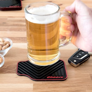 May include: Black and red rubber coaster with the word "Cheers" printed in red. A glass of beer is sitting on the coaster.