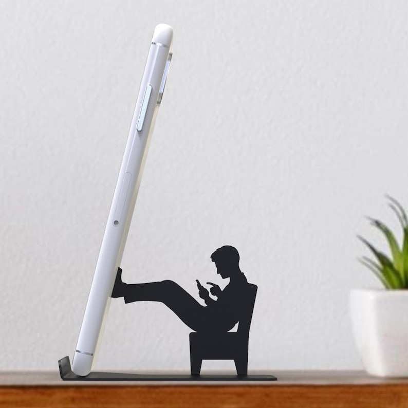 Peut inclure: Support de t&eacute;l&eacute;phone en m&eacute;tal noir en forme de personne assise sur une chaise et regardant un t&eacute;l&eacute;phone. Le t&eacute;l&eacute;phone est cal&eacute; contre la jambe de la personne.