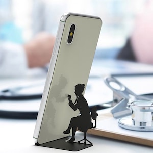 Peut inclure: Un smartphone blanc avec une silhouette noire d'une personne assise sur une chaise. La personne tient un st&eacute;thoscope. Le t&eacute;l&eacute;phone est pos&eacute; sur un support noir.
