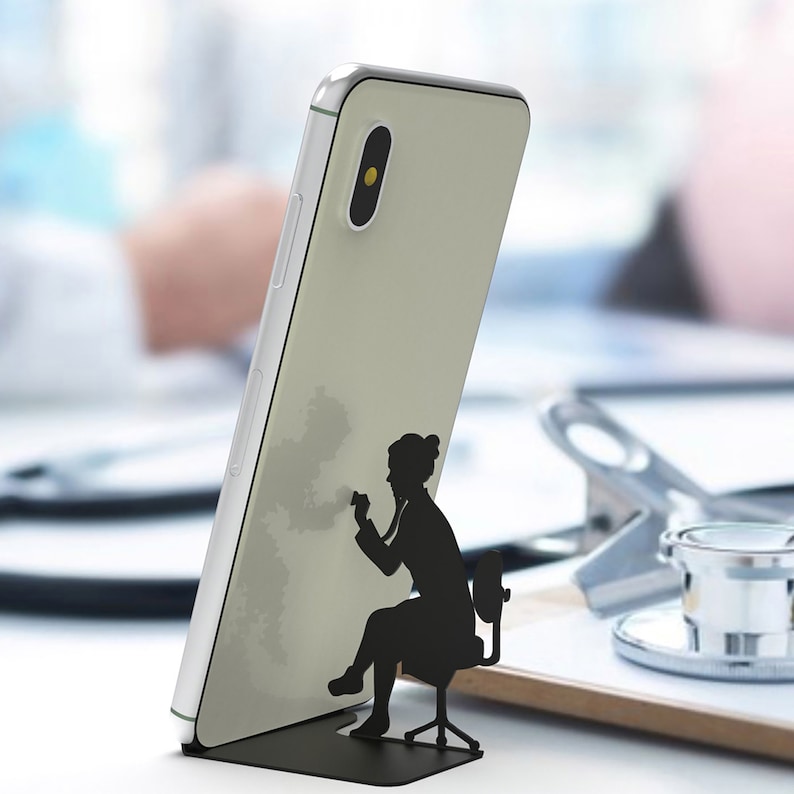 Peut inclure: Un smartphone blanc avec une silhouette noire d'une personne assise sur une chaise. La personne tient un st&eacute;thoscope. Le t&eacute;l&eacute;phone est pos&eacute; sur un support noir.