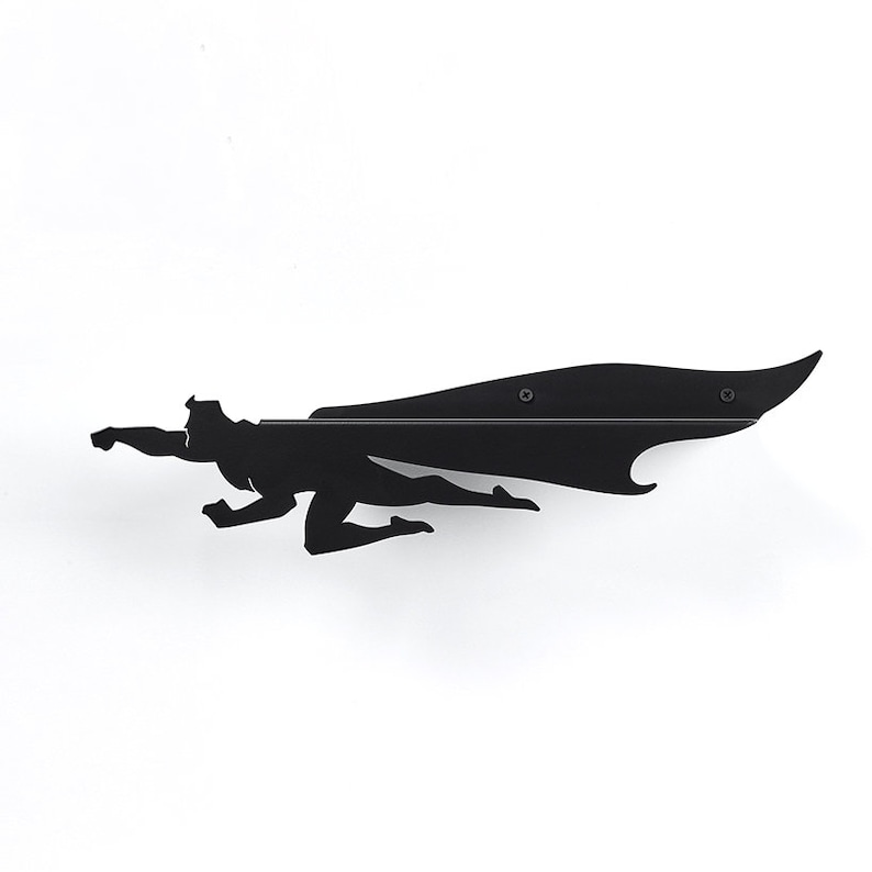Op de afbeelding: Zwart metalen wandkunst die een superheld in vlucht afbeeldt, met een lange cape. Het silhouet is op een witte muur gemonteerd. Het kunstwerk is een gestileerde weergave van een vliegende figuur, perfect voor een kinderkamer.