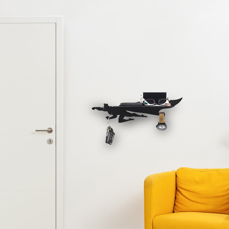 Op de afbeelding: Zwart metalen wandrek in de vorm van een superheld in vlucht. Het rek bevat een telefoon, een bril en een horloge. Sleutels hangen aan de hand van de superheld. Het rek is bevestigd aan een witte muur.