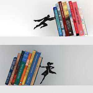 May include: Black metal bookends shaped like a superhero and a superwoman holding up books on a white shelf. The superhero is holding up a stack of books with titles like "Urban Theory", "Manhattan Voyagers", and "Jean Rhys Wide Sargasso Sea". The superwoman is holding up a stack of books with titles like "The Arabian Nights", "The Great Gatsby", and "Perfume".