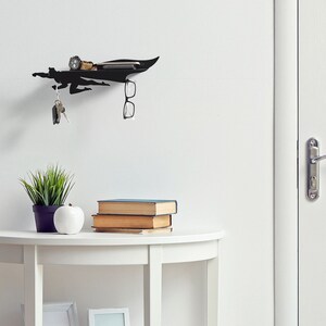 Op de afbeelding: Zwart metalen wandrek in superheldenvorm met een plank voor kleine spullen, een sleutelhouder en een brilhouder. Het rek is aan een witte muur bevestigd. Een horloge, telefoon en sleutels liggen op de plank.