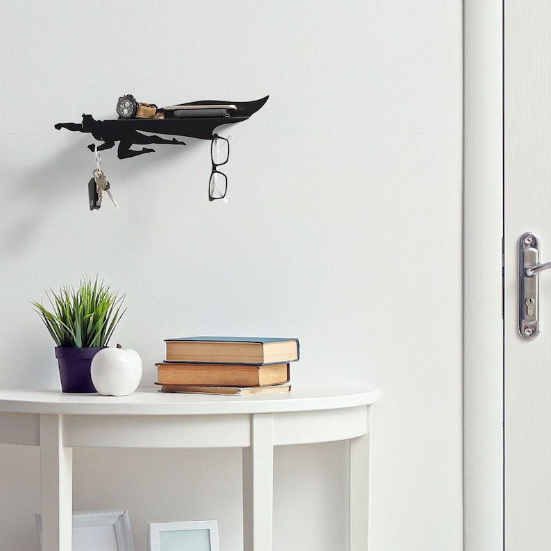 Op de afbeelding: Zwart metalen wandrek in superheldenvorm met een plank voor kleine spullen, een sleutelhouder en een brilhouder. Het rek is aan een witte muur bevestigd. Een horloge, telefoon en sleutels liggen op de plank.