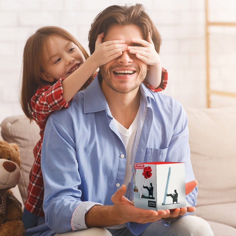 Peut inclure: Un homme sourit tandis qu'un enfant lui couvre les yeux avec ses mains. L'homme tient une petite bo&icirc;te avec une fleur rouge et une silhouette d'une personne sur une &eacute;chelle sur le devant. La bo&icirc;te dit "Art Deco"