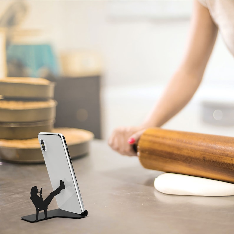 Peut inclure: Un support de t&eacute;l&eacute;phone en m&eacute;tal noir avec une silhouette d'une personne assise les jambes en l'air. Le support tient un smartphone blanc.