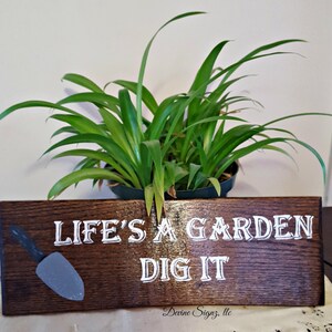 K&ouml;nnte beinhalten: Ein dunkelbraunes Holzschild mit wei&szlig;er Aufschrift "Life's a Garden Dig It". Auf der linken Seite des Schildes ist eine kleine graue Schaufel gemalt. Das Schild steht auf einer wei&szlig;en Oberfl&auml;che vor einer gr&uuml;nen Pflanze.