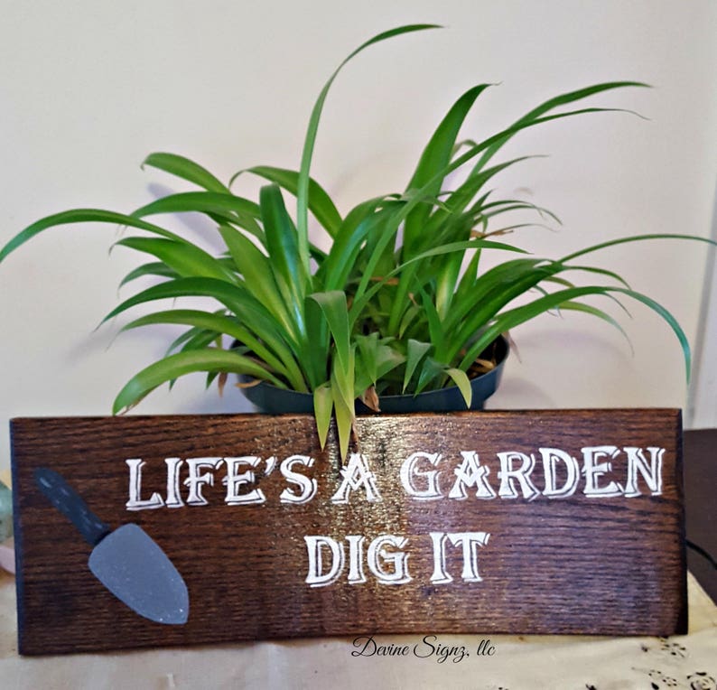 K&ouml;nnte beinhalten: Ein dunkelbraunes Holzschild mit wei&szlig;er Aufschrift "Life's a Garden Dig It". Auf der linken Seite des Schildes ist eine kleine graue Schaufel gemalt. Das Schild steht auf einer wei&szlig;en Oberfl&auml;che vor einer gr&uuml;nen Pflanze.