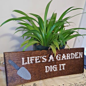 K&ouml;nnte beinhalten: Ein Holzschild mit dem Text "Life's a garden dig it" in wei&szlig;er Farbe. Links auf dem Schild befindet sich eine grau bemalte Schaufel. Das Schild steht auf einem Tisch mit einer Tischdecke mit Blumenmuster.