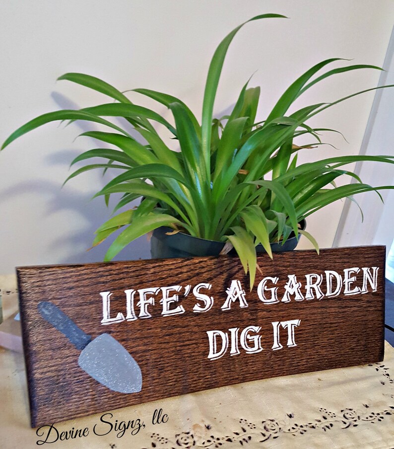 K&ouml;nnte beinhalten: Ein Holzschild mit dem Text "Life's a garden dig it" in wei&szlig;er Farbe. Links auf dem Schild befindet sich eine grau bemalte Schaufel. Das Schild steht auf einem Tisch mit einer Tischdecke mit Blumenmuster.