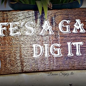 K&ouml;nnte beinhalten: Ein Holzschild mit dem Text "Life's a Garden Dig It" in wei&szlig;en Buchstaben. Auf der linken Seite des Schildes ist eine kleine silberne Schaufel gemalt.