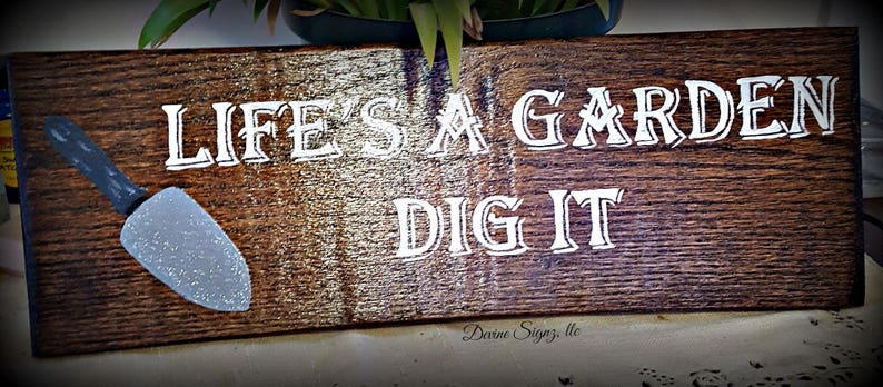 K&ouml;nnte beinhalten: Ein Holzschild mit dem Text "Life's a Garden Dig It" in wei&szlig;en Buchstaben. Auf der linken Seite des Schildes ist eine kleine silberne Schaufel gemalt.