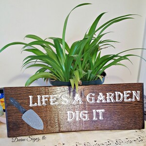 K&ouml;nnte beinhalten: Ein Holzschild mit dem Text "Life's a garden, dig it" in wei&szlig;en Buchstaben. Auf der linken Seite des Schildes ist eine kleine graue Schaufel gemalt.