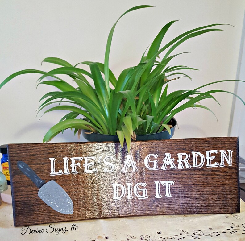 K&ouml;nnte beinhalten: Ein Holzschild mit dem Text "Life's a garden, dig it" in wei&szlig;en Buchstaben. Auf der linken Seite des Schildes ist eine kleine graue Schaufel gemalt.