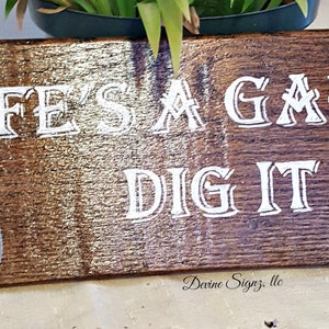 K&ouml;nnte beinhalten: Ein Holzschild mit dem Text "Life's a Garden Dig It" in wei&szlig;en Buchstaben. Auf der linken Seite des Schildes ist eine kleine silberne Schaufel gemalt.