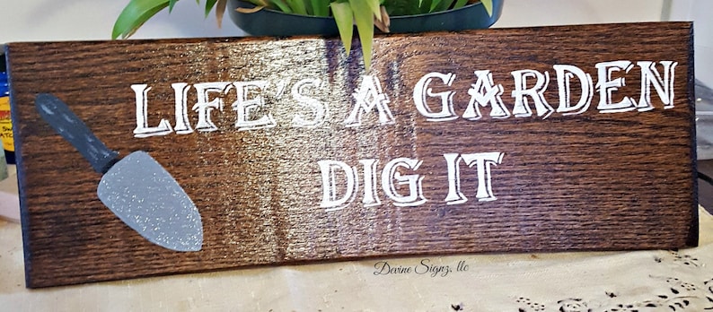 K&ouml;nnte beinhalten: Ein Holzschild mit dem Text "Life's a Garden Dig It" in wei&szlig;en Buchstaben. Auf der linken Seite des Schildes ist eine kleine silberne Schaufel gemalt.