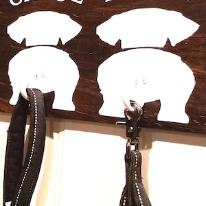 May include: A wooden dog leash holder with two white dog silhouettes. The silhouettes have the names "Chloe" and "Abby" written above them. Two black leashes are hanging from the hooks.