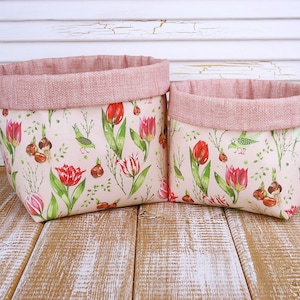 Puede incluir: Dos cestas de almacenamiento de tela. Las cestas presentan un estampado floral con tulipanes rojos, hojas verdes y pequeños pájaros sobre un fondo rosa claro. Los bordes superiores están forrados con una tela rosa texturizada. Las cestas están sobre una superficie de madera.