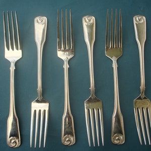 May include: Six silver forks with ornate handles featuring a rose design. The forks are arranged in a row on a blue surface.