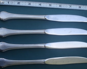 May include: Four silver butter knives with a decorative handle design. The knives are approximately 7 inches long, with a curved blade and a handle that widens at the end. The knives are arranged on a dark blue surface.
