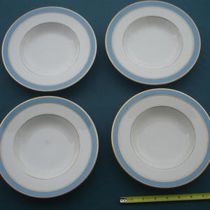 May include: Four white ceramic plates with a light blue border and a gold trim. The plates are arranged in a square formation. A measuring tape is at the bottom of the image.
