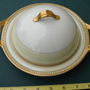 May include: A white and gold porcelain butter dish with a lid and handles. The dish has a gold rim and a gold handle on the lid. The base is a pale yellow color with a gold trim. The dish is approximately 8 inches in diameter.