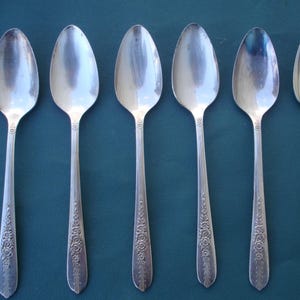 May include: Six silver teaspoons with ornate floral designs on the handles. The spoons are arranged in a row on a blue surface.