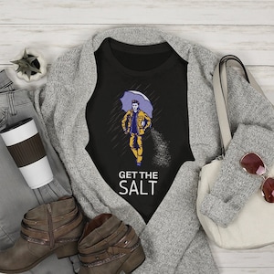 May include: Black t-shirt with a graphic of a person walking in the rain with an umbrella and the text "Get the Salt". The t-shirt is on a gray cardigan with a pair of brown ankle boots, a pair of jeans, a coffee cup, and a white handbag.