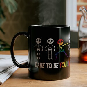 May include: Black coffee mug with a white design of four skeletons. The skeletons are in a line and the one on the right is doing a dab. The text "Dare to be yourself" is below the skeletons.