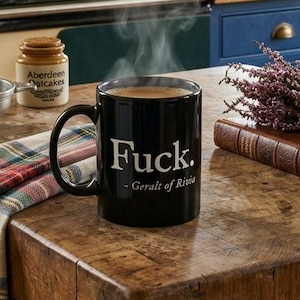 May include: A black mug with the word "Fuck" in white, with steam rising from the coffee. The mug sits on a wooden countertop with a plaid cloth, a book, and a bundle of lavender. The kitchen has green cabinets and a stove.