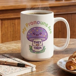 May include: A white ceramic mug featuring a colorful graphic of a smiling purple mushroom with polka dots. The text "My Pronouns Are" arches above the mushroom, while the words "THEY THEM" are printed on the mushroom stem with a small heart.