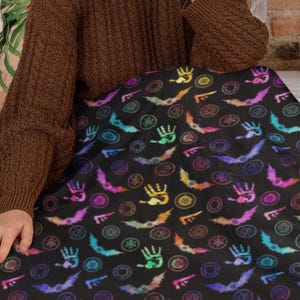 May include: A black blanket with a repeating pattern of colorful bats, hands, and other symbols. The blanket is spread out, covering a bed. The colors include purple, blue, orange, and yellow. The blanket is made of a soft material.