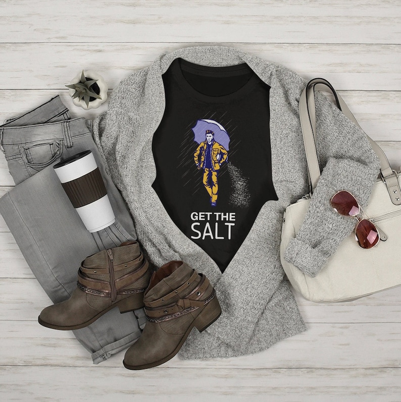 May include: Black t-shirt with a graphic of a person walking in the rain with an umbrella and the text "Get the Salt". The t-shirt is on a gray cardigan with a pair of brown ankle boots, a pair of jeans, a coffee cup, and a white handbag.