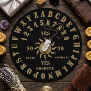May include: A circular black spirit board featuring gold lettering of the alphabet, numbers zero through nine, and the words YES, NO, MAYBE, REPHRASE, and GOODBYE. A clear crystal point pendant on a gold chain rests in the center over a sun icon.