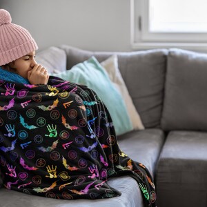May include: A black throw blanket with a colorful pattern of bats, hands, and other symbols. The blanket is draped over a person wearing a pink beanie and a blue-lined hood, sitting on a gray couch.
