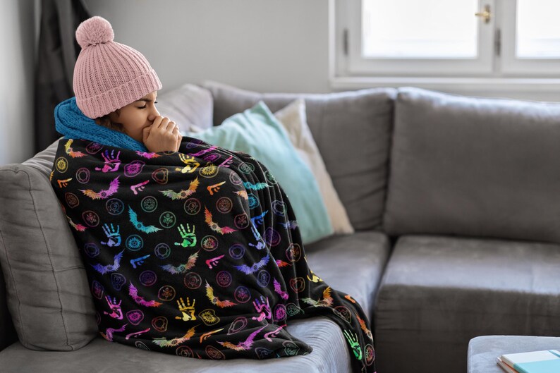 May include: A black throw blanket with a colorful pattern of bats, hands, and other symbols. The blanket is draped over a person wearing a pink beanie and a blue-lined hood, sitting on a gray couch.