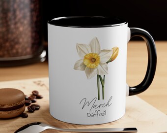 May include: White ceramic mug with a black rim and handle. The mug features a white daffodil with a yellow center and the text "March Daffodil".