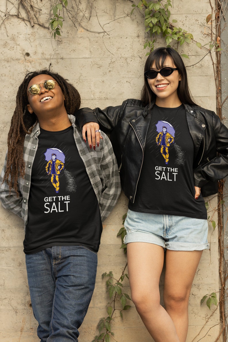 May include: Two people wearing black t-shirts with a graphic of a person holding an umbrella and the text "Get the Salt". The person on the left is wearing a plaid shirt and jeans. The person on the right is wearing a black leather jacket and denim shorts.