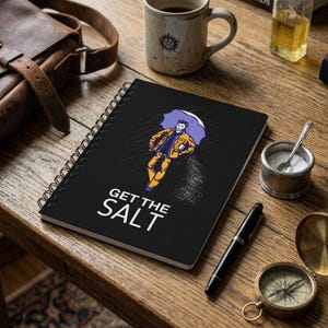 May include: A black spiral-bound notebook featuring a graphic illustration of a person in a yellow jacket holding a purple umbrella while sprinkling salt. The text GET THE SALT is printed in white capital letters at the bottom of the cover.