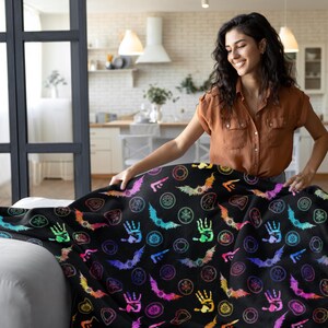 May include: A black blanket with a colorful Halloween-themed pattern featuring bats, hands, and circular designs. The blanket is spread out on a white couch. The person in the image is wearing a brown shirt.