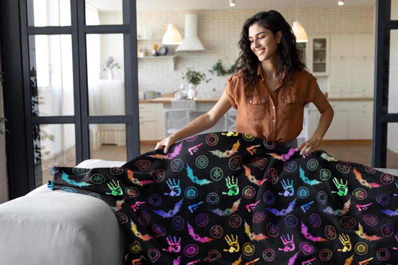 May include: A black blanket with a colorful Halloween-themed pattern featuring bats, hands, and circular designs. The blanket is spread out on a white couch. The person in the image is wearing a brown shirt.