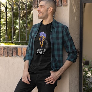 May include: A man wearing a black t-shirt with a graphic of a person holding an umbrella and the text "GET THE SALT". He is also wearing a green and black plaid shirt and black jeans.