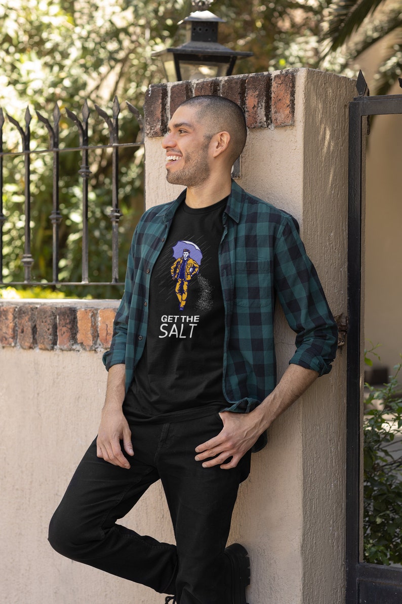 May include: A man wearing a black t-shirt with a graphic of a person holding an umbrella and the text "GET THE SALT". He is also wearing a green and black plaid shirt and black jeans.