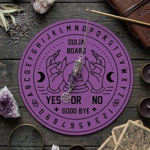 May include: A round purple Ouija board featuring black printed letters, numbers, and illustrations of hands holding a crystal ball. A silver-toned metal pendulum with a pointed amethyst crystal and a pentagram charm hangs over the board.