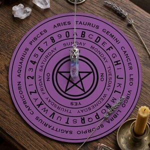 May include: A circular purple dowsing board featuring black text including the alphabet, numbers 0-9, days of the week, zodiac signs, and yes or no options. A silver-toned metal chain holds a multi-colored crystal pendulum over a central pentagram.