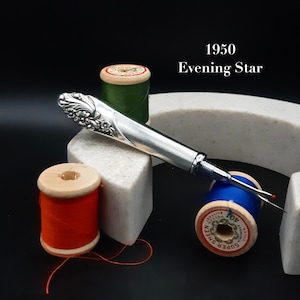 Peut inclure: Un coupe-fil en argent avec un motif floral sur la poignée. Le coupe-fil est posé sur une surface blanche avec des bobines de fil rouge, vert et bleu.