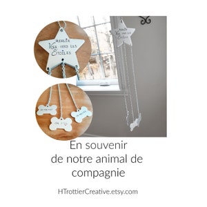 May include: A silver wind chime with a star-shaped top and three bone-shaped charms. The star has the text "MERLIN VOLE VERS LES ETOILES" engraved on it. The bone charms have the dates "01 06-07", "24+06-10", and "ON T'" engraved on them. The wind chime is hanging in a window. The text "En souvenir de notre animal de compagnie HTrottierCreative.etsy.com" is at the bottom of the image.