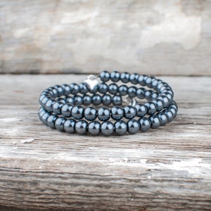 May include: A dark gray pearl bracelet with a silver heart charm. The bracelet is made of multiple strands of pearls and is coiled on a wooden surface.